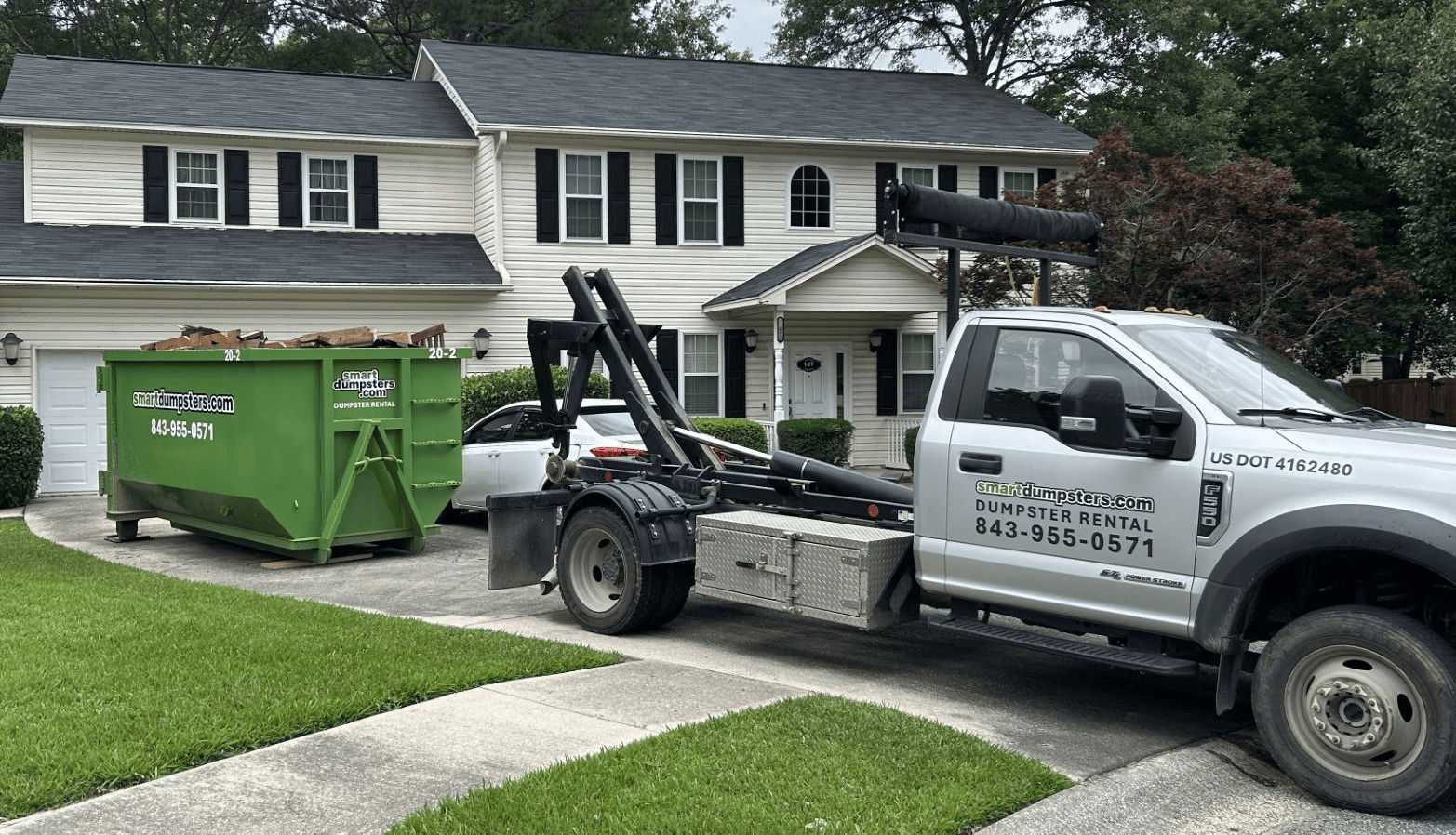 A dumpster rental Charleston truck is parked in the driveway of a suburban house in Berkeley, Dorchester County. A green dumpster filled with debris sits on the well-maintained lawn beside the white home with black shutters.