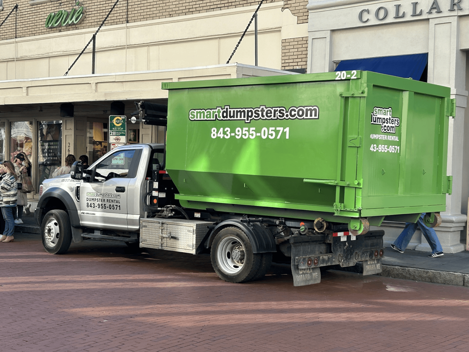 A silver truck with a bright green dumpster, advertising smartdumpsters.com and 843-955-0571, is parked on a brick street. People walk nearby. Perfect for those seeking dumpster rental Charleston, SC or in Dorchester County.