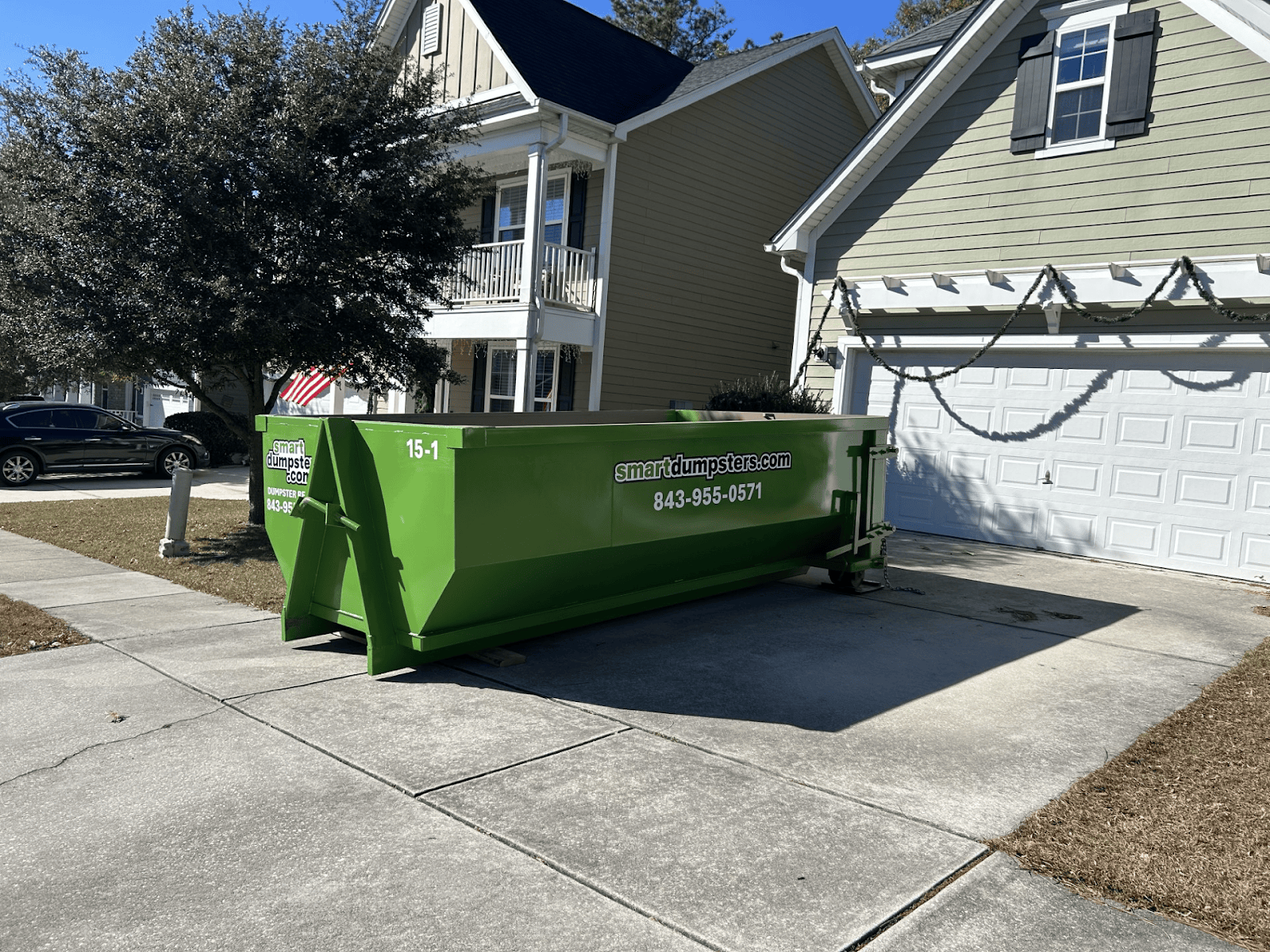 A large green dumpster rental Charleston sits on the driveway in front of a suburban house with white trim and a double garage. There are leafless trees, a car, and an American flag in the background, evoking scenes from Berkeley or Dorchester County.