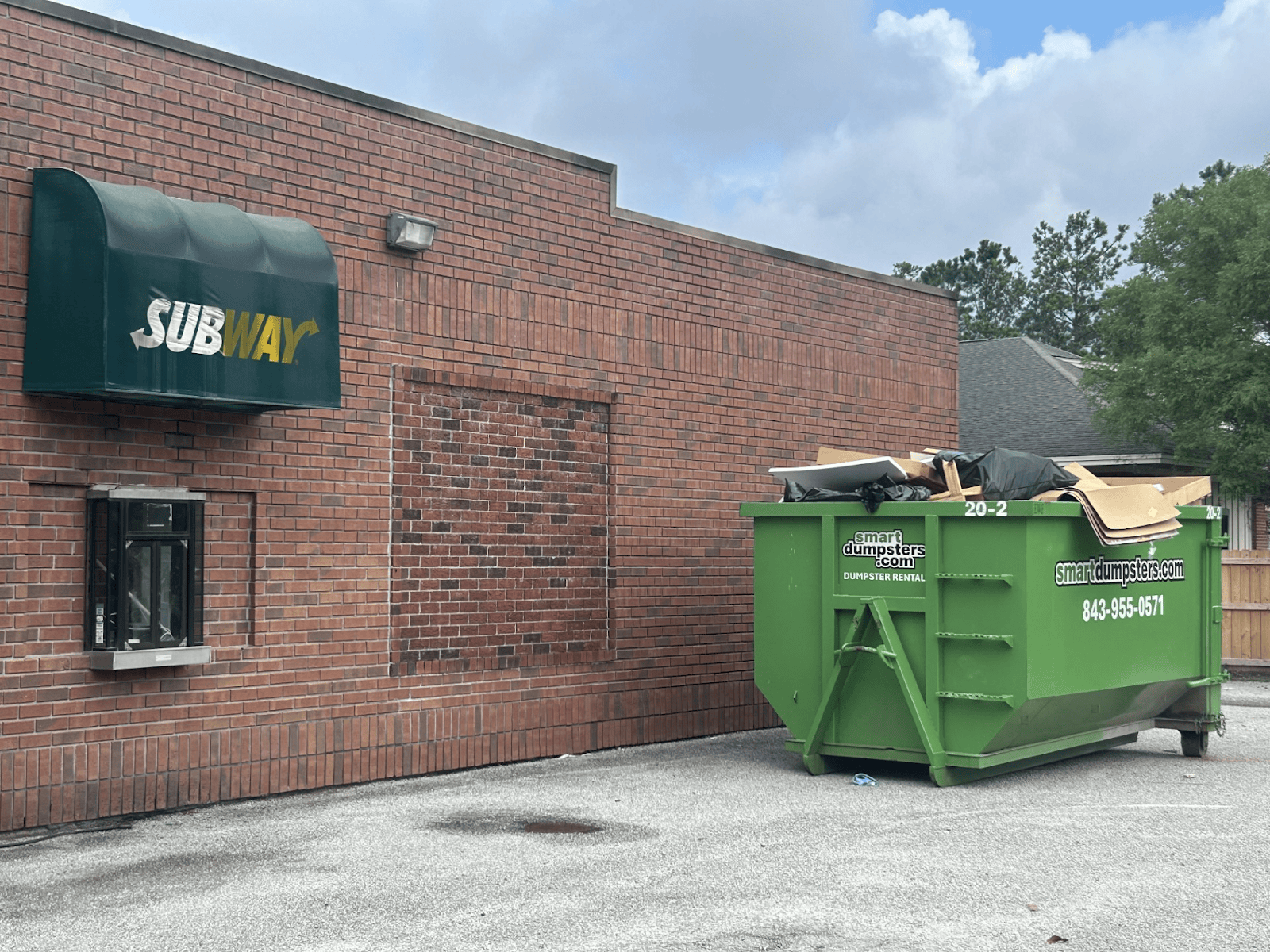 A green dumpster rental Charleston sits filled with debris in a parking lot next to a brick Subway restaurant with a drive-thru window and green awning. Trees and houses are visible in the Berkeley, SC background.