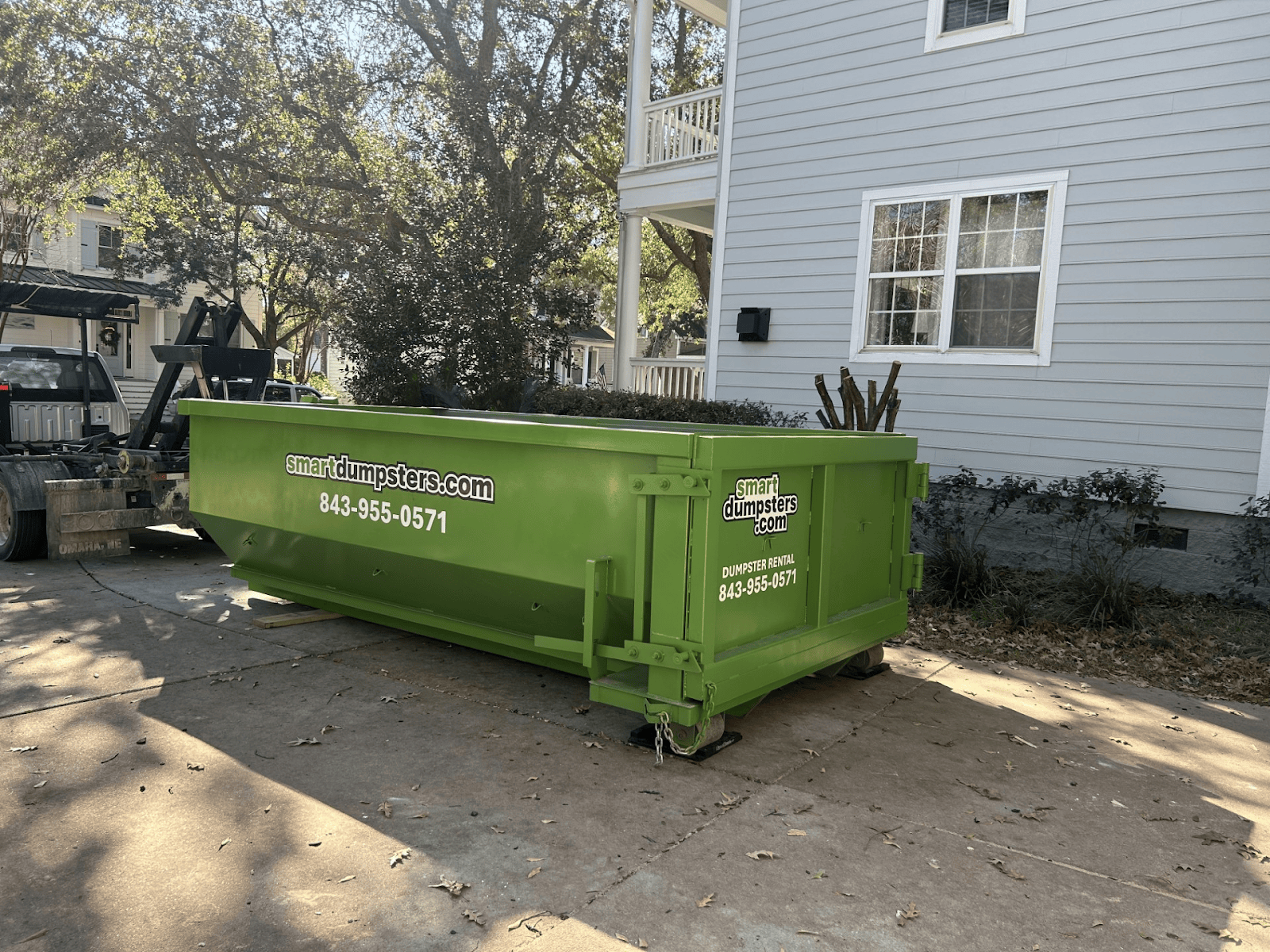 A bright green dumpster labeled "smartdumpsters.com" with a phone number, ideal for dumpster rental Charleston, sits on a driveway beside a light gray house with white trim and several windows. Trees cast shadows on the ground.