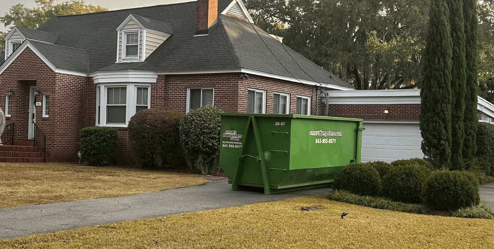 A large green dumpster sits on the driveway in front of a brick house with a white garage and well-kept shrubs, under an overcast sky in Dorchester County.