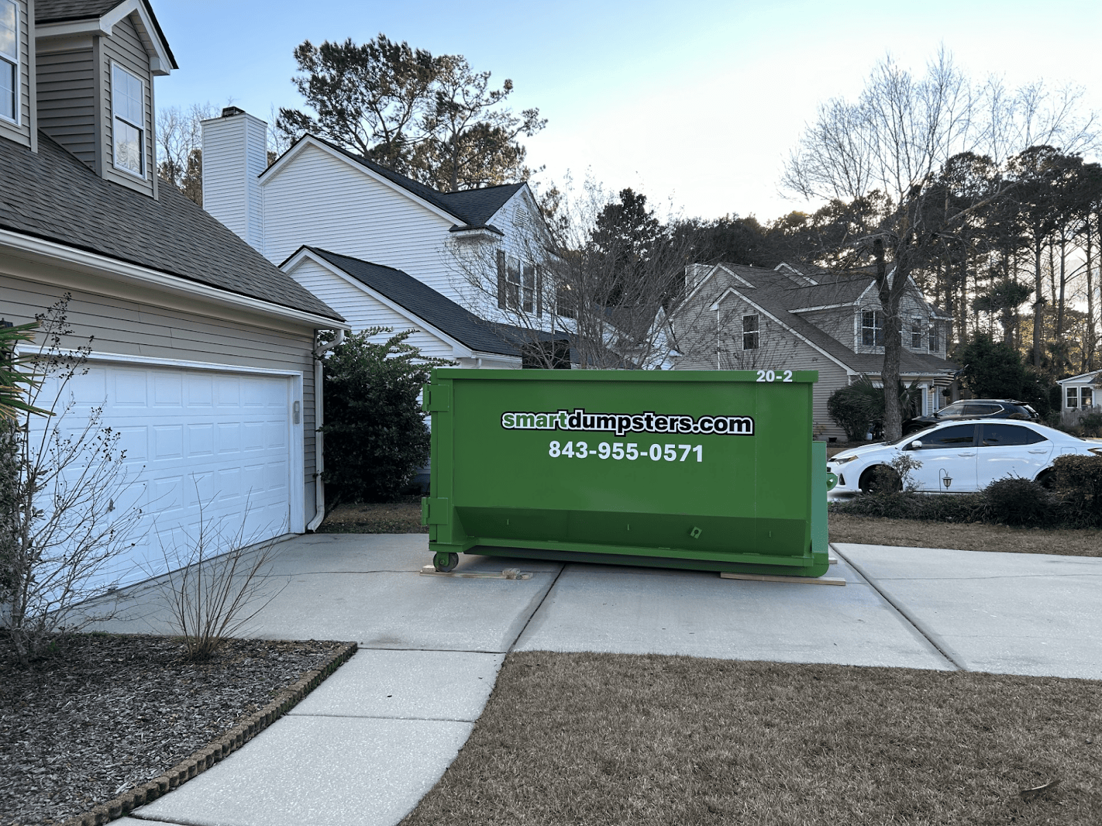 A large green dumpster labeled "smartdumpsters.com 843-955-0571" sits in a residential driveway in Berkeley, SC, in front of a two-car garage, surrounded by houses, trees, and parked cars.