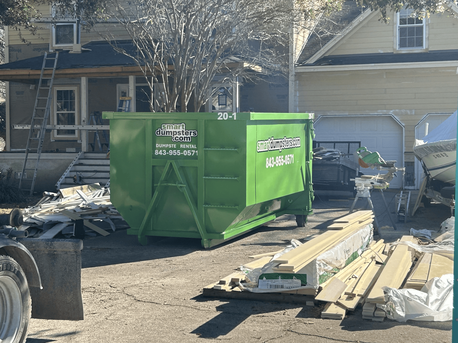A large green dumpster labeled "smartdumpsters.com" sits in a driveway in front of a house under renovation in Dorchester County, surrounded by construction materials, debris, and equipment—perfect for your dumpster rental Charleston, SC needs.