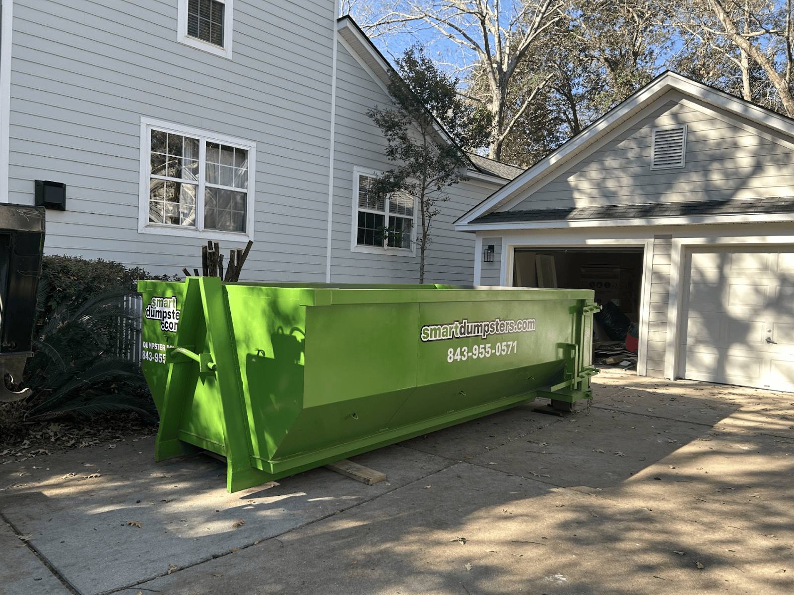 A large, bright green dumpster labeled "smartdumpsters.com 843-955-0571" sits on the driveway outside a light gray house with a two-car garage on a sunny day, perfect for your dumpster rental Charleston, SC or Dorchester County needs.