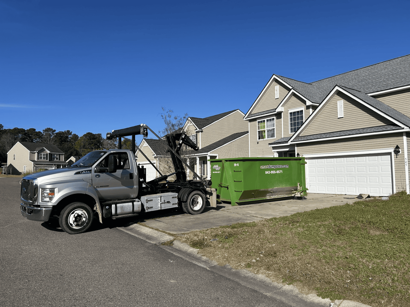 A silver truck from a dumpster rental Charleston service is delivering a large green dumpster to the driveway of a suburban house in Berkeley or Dorchester County on a sunny day. The house features beige siding, white trim, and a two-car garage.