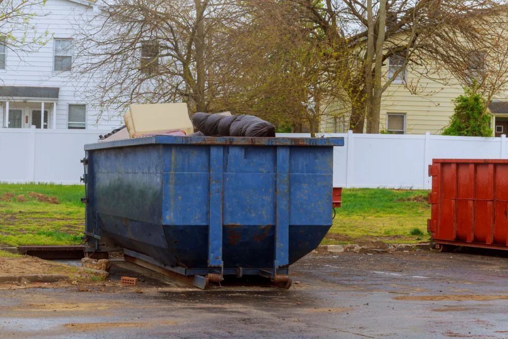 A large blue dumpster with trash bags and boxes sits near houses and a white fence outdoors.