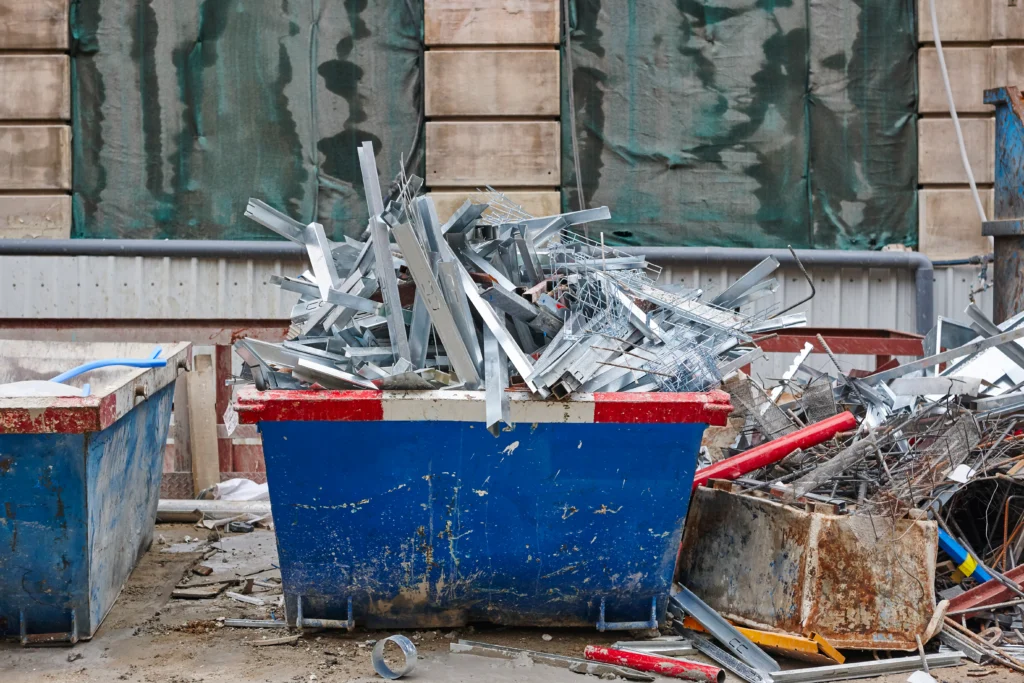 A blue dumpster filled with scrap metal stands by a rusty bin at a construction site during the day.